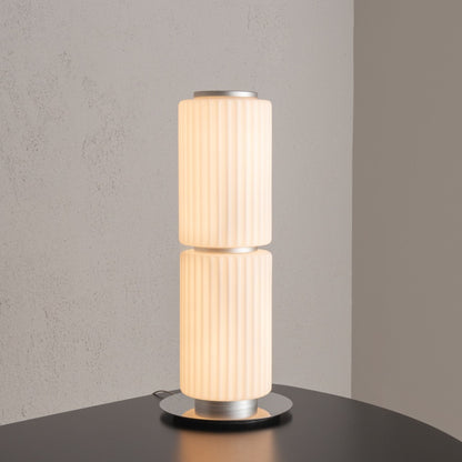 Tall cylindrical table lamp with ribbed white glass and a round metal base placed on a black surface.