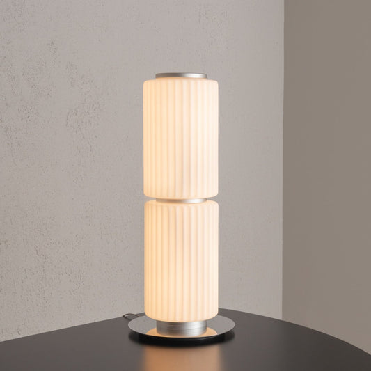 Tall cylindrical table lamp with ribbed white glass and a round metal base placed on a black surface.