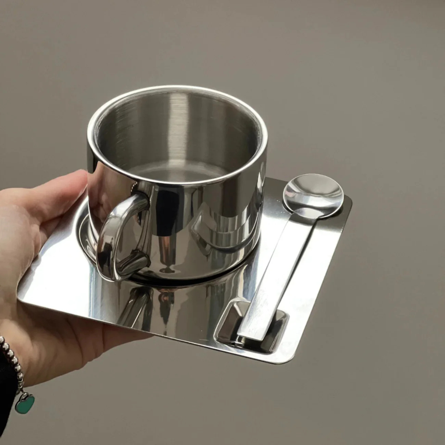 Hand holding a stainless steel cup placed on a square reflective saucer with a matching metal spoon.