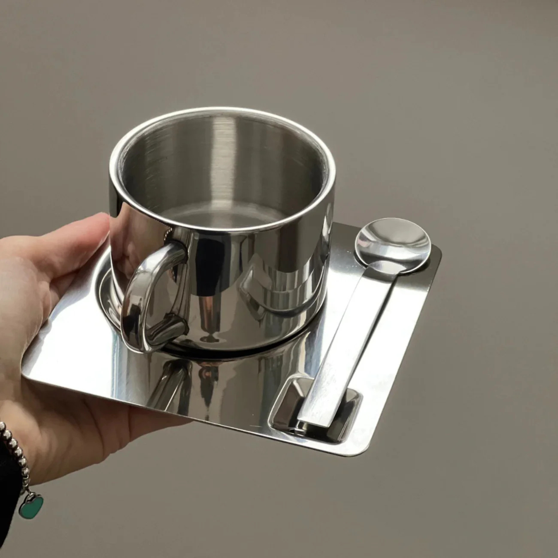 Hand holding a stainless steel cup placed on a square reflective saucer with a matching metal spoon.