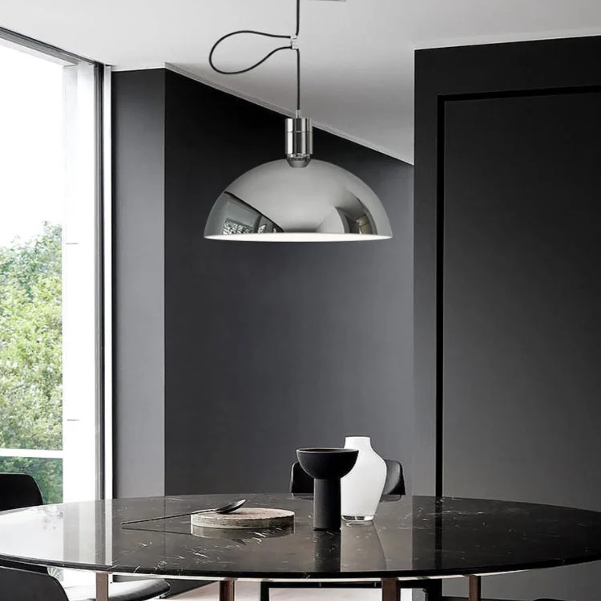 A shiny chrome dome pendant lamp hanging above a dark marble dining table in a modern interior with dark walls.