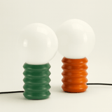 Two light bulbs with ribbed ceramic bases, one green and one orange, on a white background.
