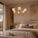 A modern six-light chandelier with a brushed gold metal frame and frosted blush pink glass shades hangs over a neatly made bed in a softly lit, elegant bedroom.