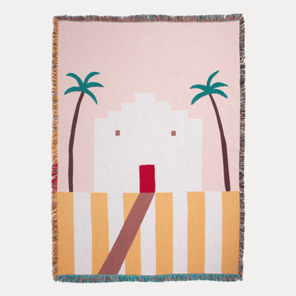 Woven blanket with a design of palm trees and a building on a pink background