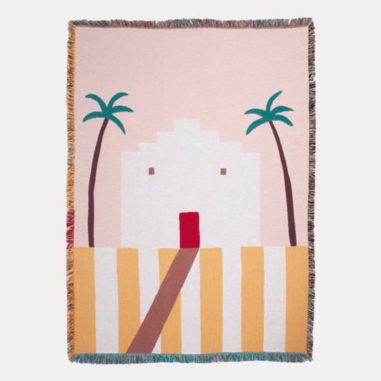 Woven blanket with a design of palm trees and a building on a pink background