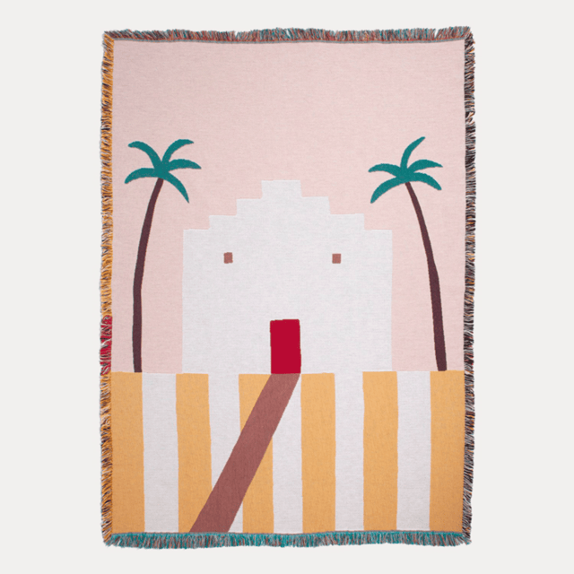 Woven blanket with a design of palm trees and a building on a pink background