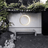 Circular light fixture on a white wall with greenery and a bench in the foreground