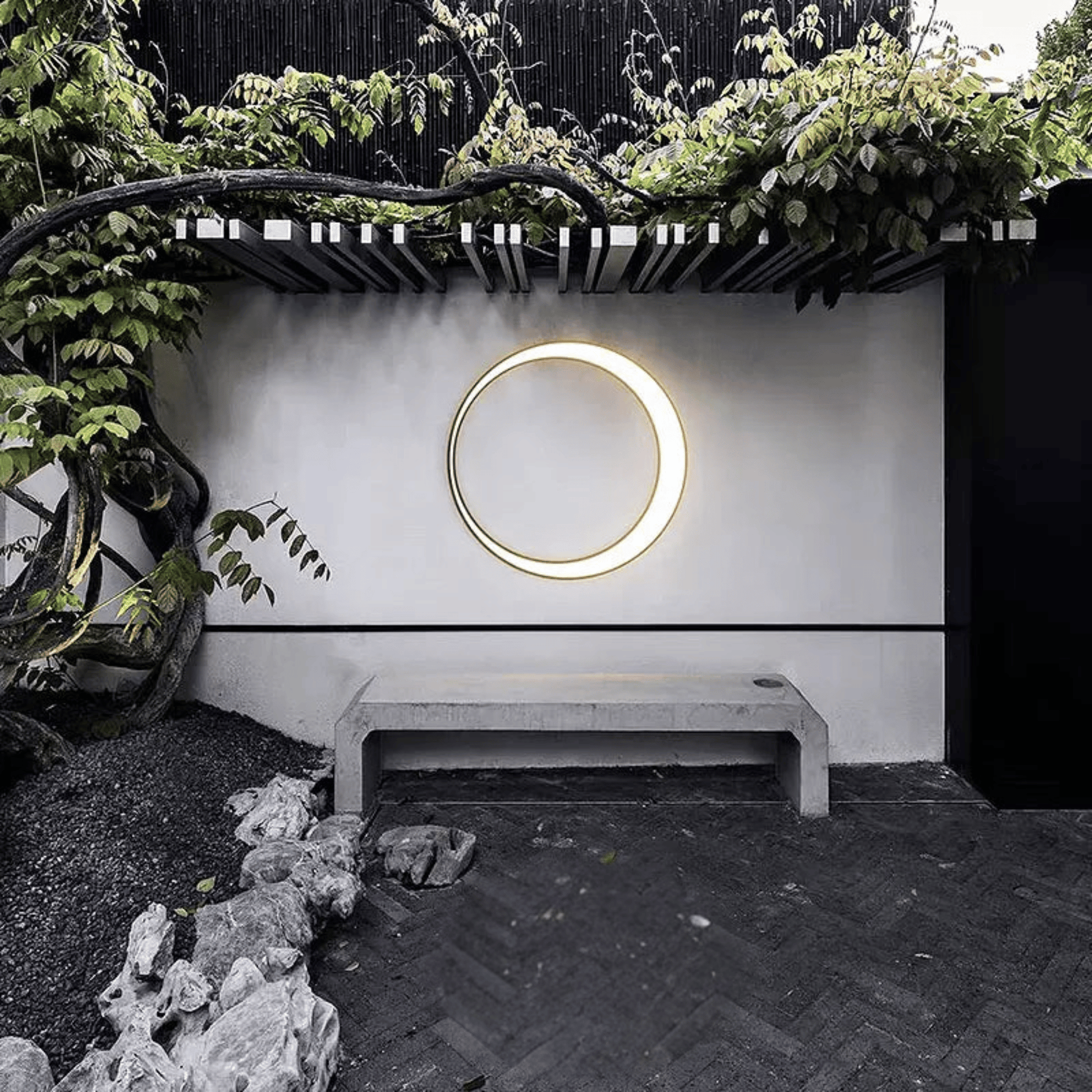 Circular light fixture on a white wall with greenery and a bench in the foreground