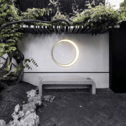 Circular light fixture on a white wall with greenery and a bench in the foreground