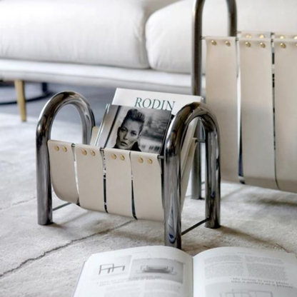 A white leather and chrome metal magazine holder sitting on a gray carpet, holding a magazine titled “RODIN,” with an open book placed nearby.