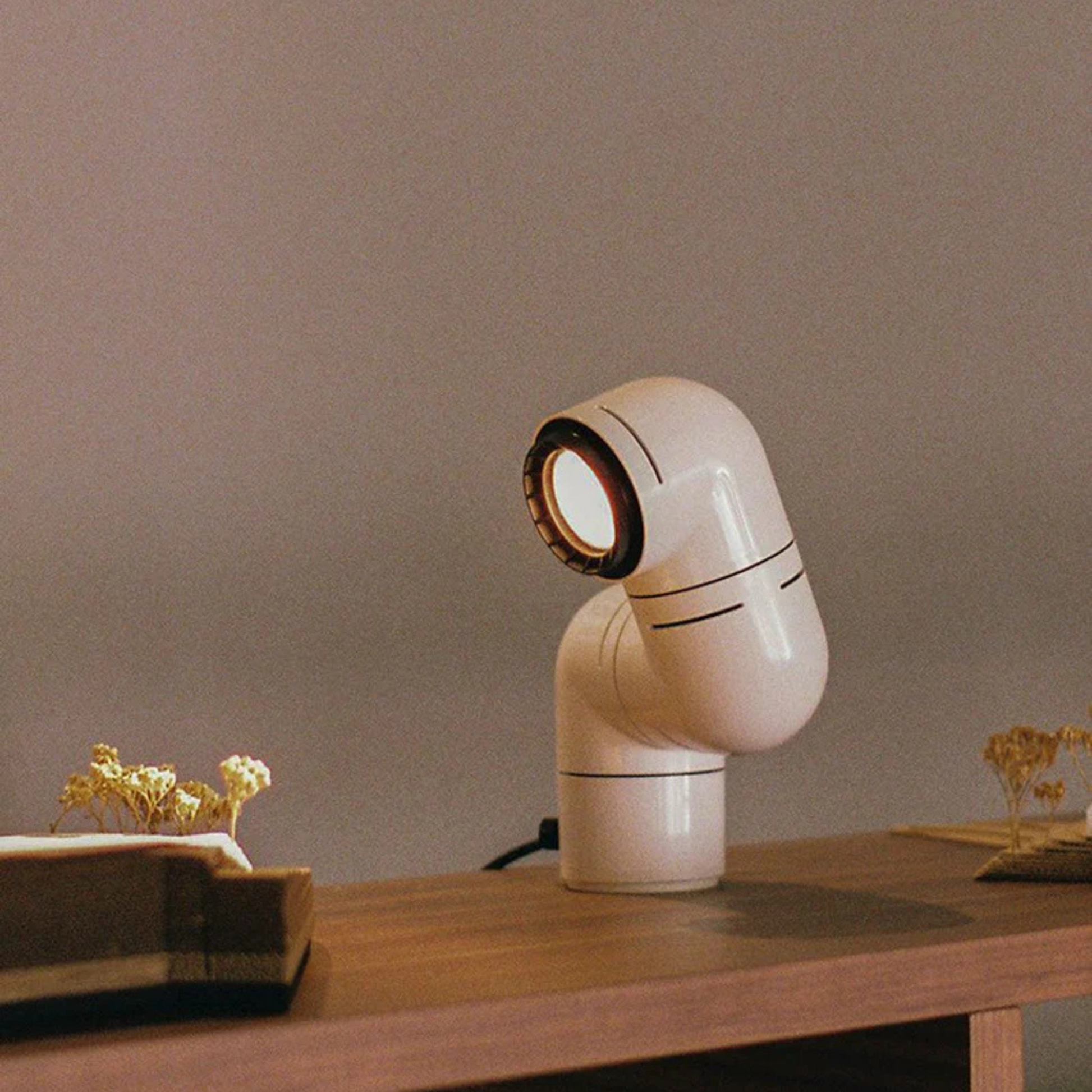A white elbow-shaped desk lamp on a wooden table with flowers and a book in a softly lit room.