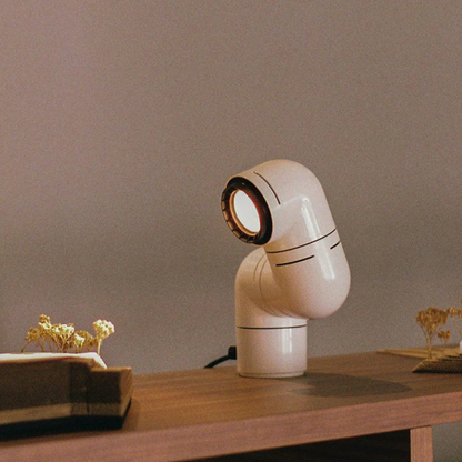 A white elbow-shaped desk lamp on a wooden table with flowers and a book in a softly lit room.