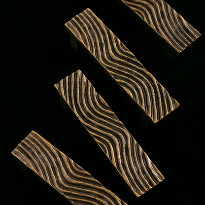 A modern wavy brass handle mounted on a matte black cabinet door, highlighting its detailed texture.