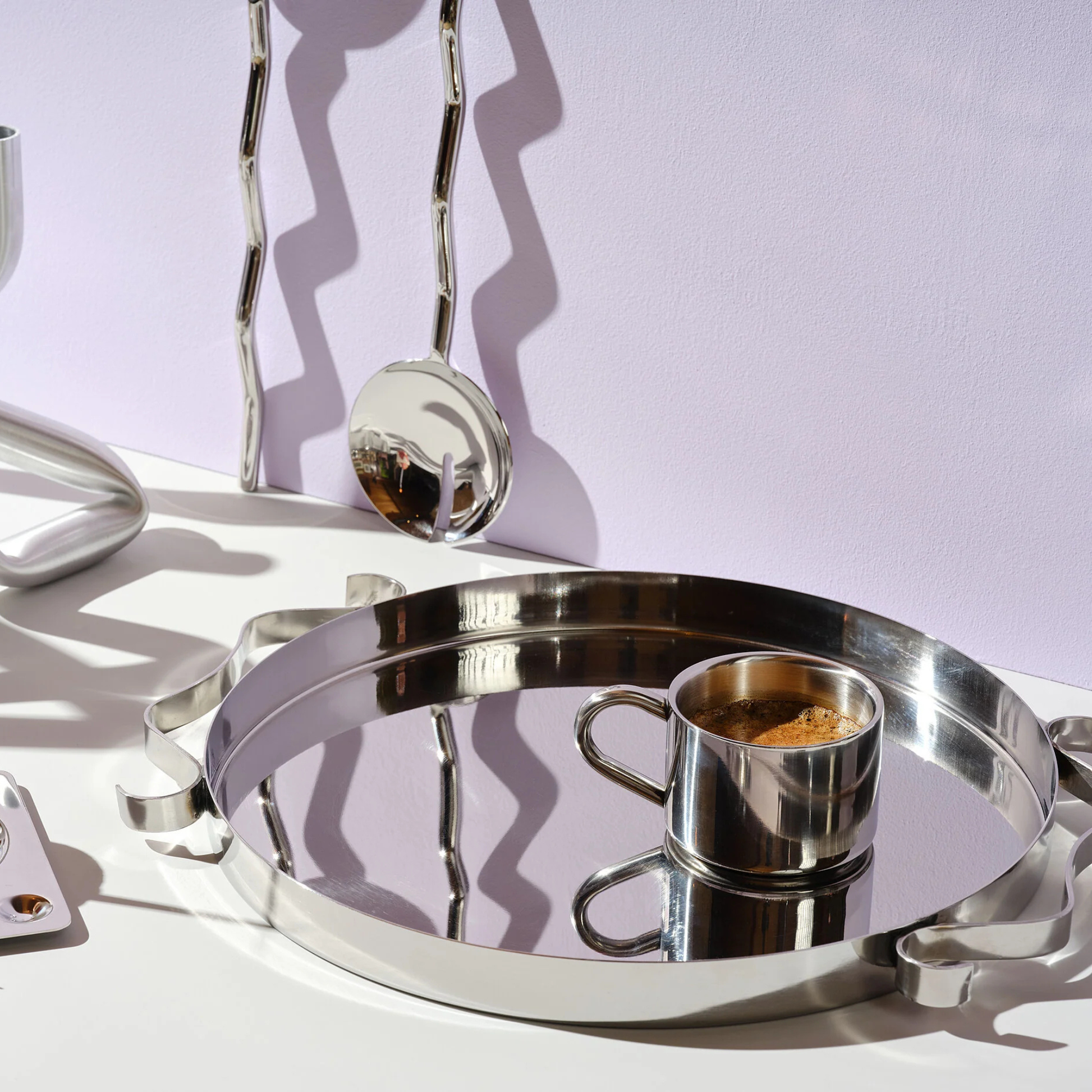 Stainless steel coffee cup on a reflective square saucer with spoon, isolated on a plain white background.