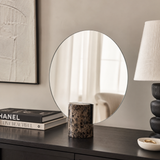 Round mirror on a dark wooden surface with books, a candle, and a lamp in a room setting.