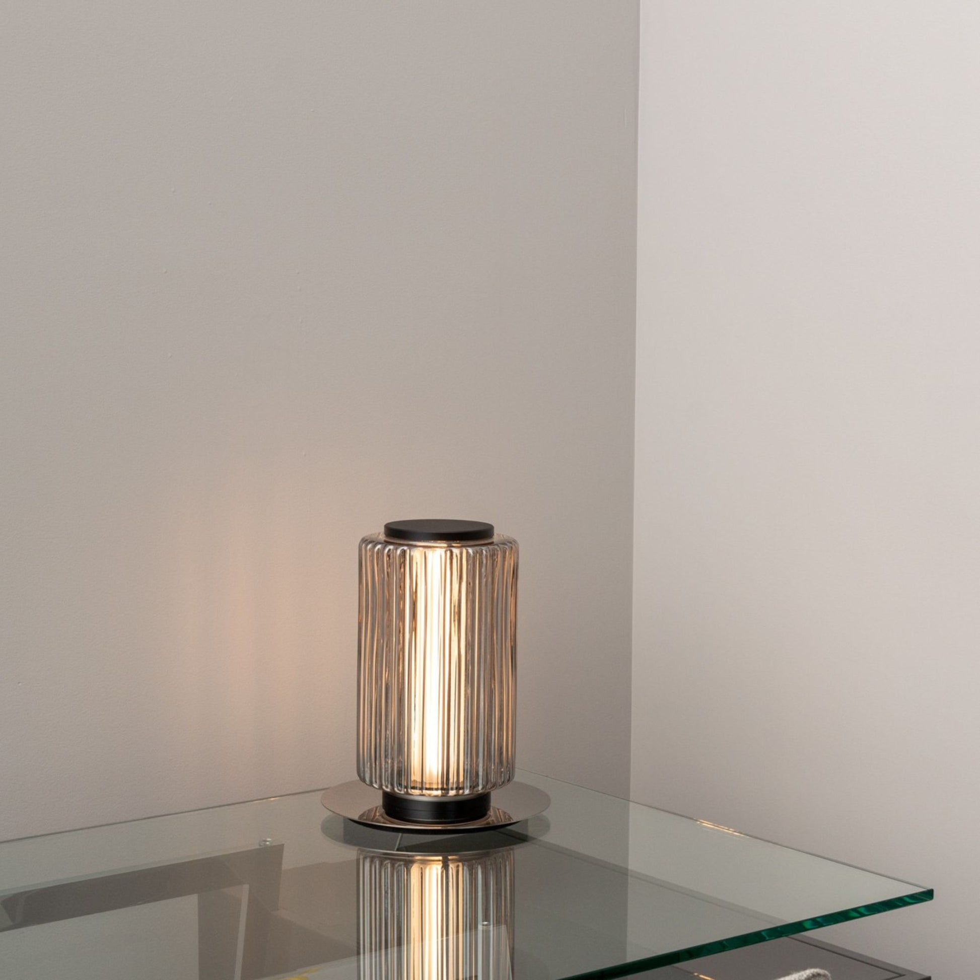 Compact ribbed-glass table lamp glowing softly on a glass side table against a light wall.