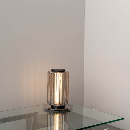 Compact ribbed-glass table lamp glowing softly on a glass side table against a light wall.