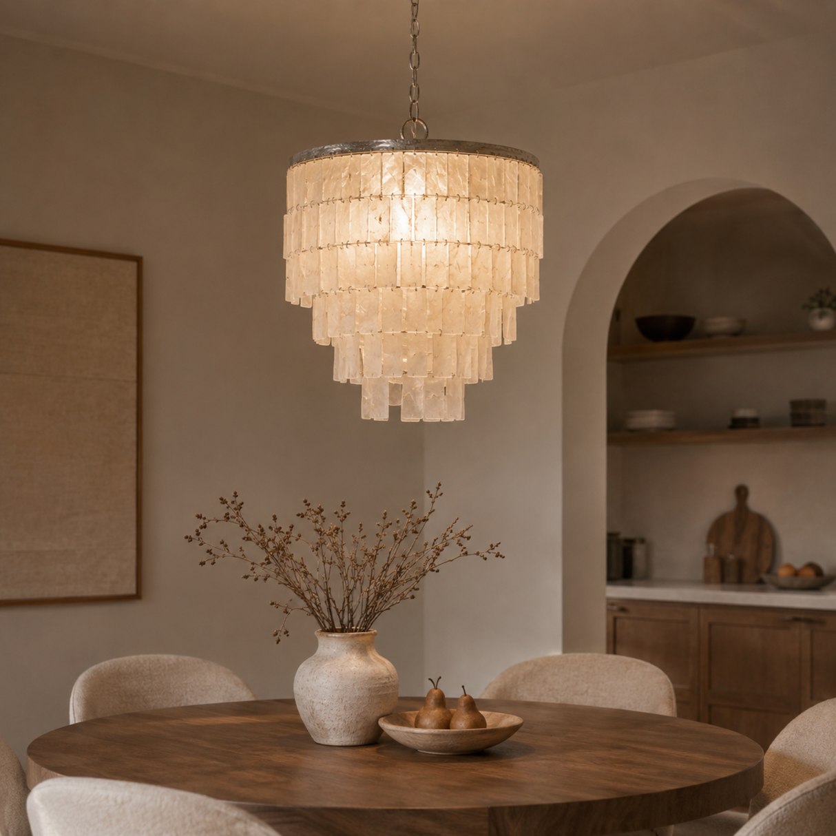 Chandelier hanging above a dining table with a vase and fruit bowl in a room with shelves and a painting.