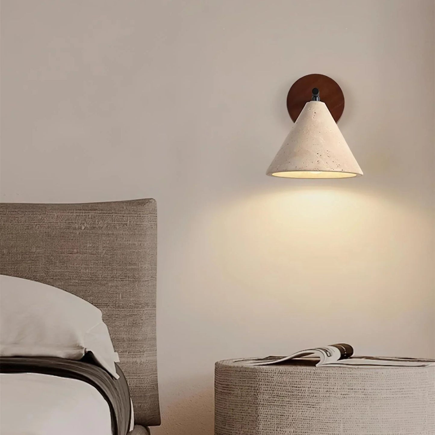 A warm, soft-glowing wall lamp with a wooden base and white shade, lighting up a minimal bedroom space with light bedding.