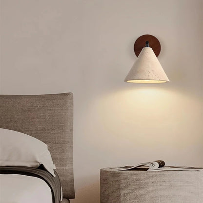 A warm, soft-glowing wall lamp with a wooden base and white shade, lighting up a minimal bedroom space with light bedding.