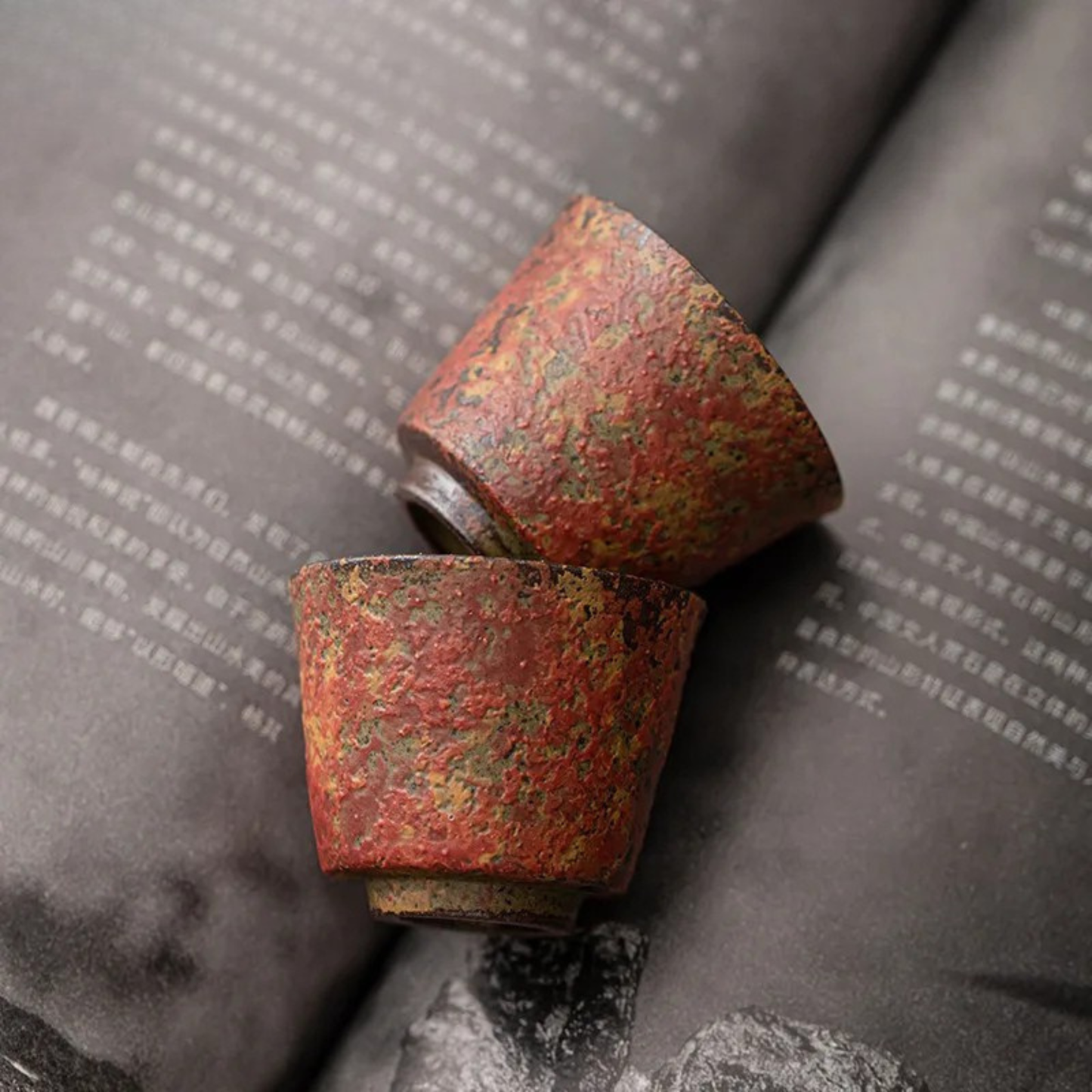 Two red textured ceramic tea cups lying on an open book with Chinese text.