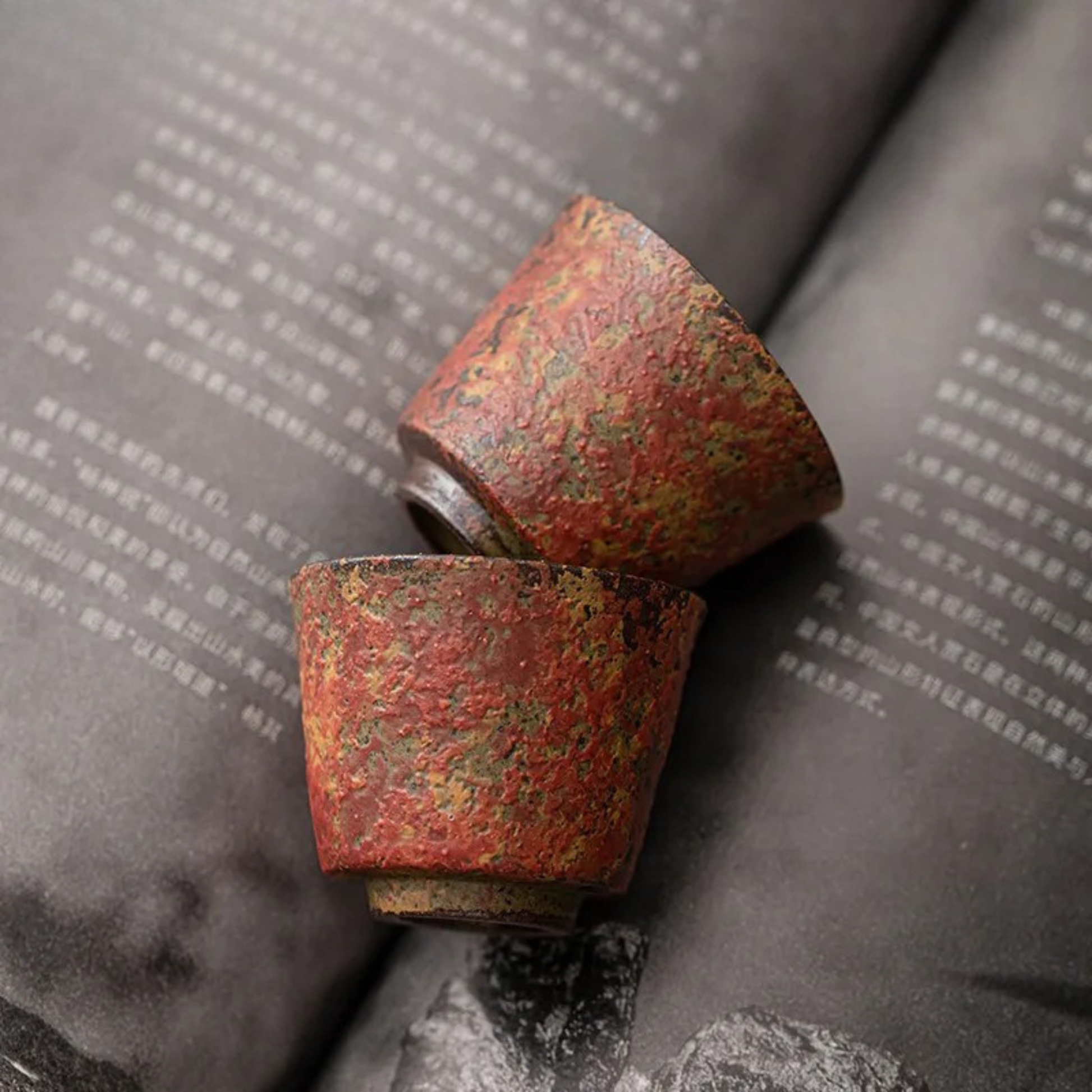 Two red textured ceramic tea cups lying on an open book with Chinese text.