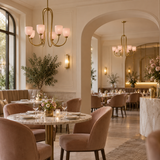 An elegant dining room interior with blush pink velvet chairs, round marble-top tables, gold chandeliers, and warm cream walls with arched alcoves.