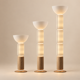Three modern floor lamps in graduated heights, featuring frosted white bowl-shaped shades, stacked translucent cylindrical sections, light natural wood bases, and warm glowing LED lighting, set against a solid warm beige background.