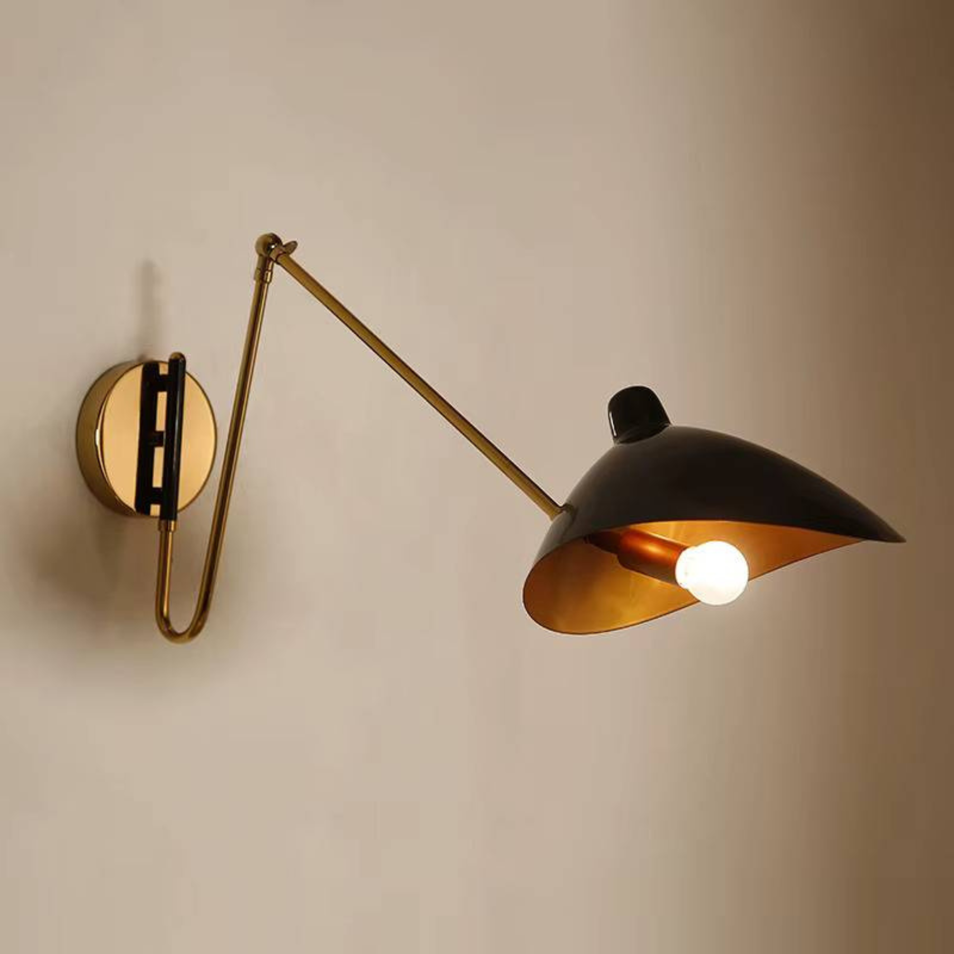 Close-up of a gold and black wall lamp with an angled shade and glowing bulb against a beige background.