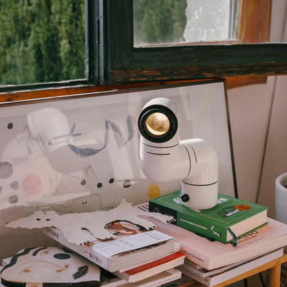 A white elbow-shaped desk lamp casting light on a stack of books, with artwork and plants in the background, creating a cozy ambiance.