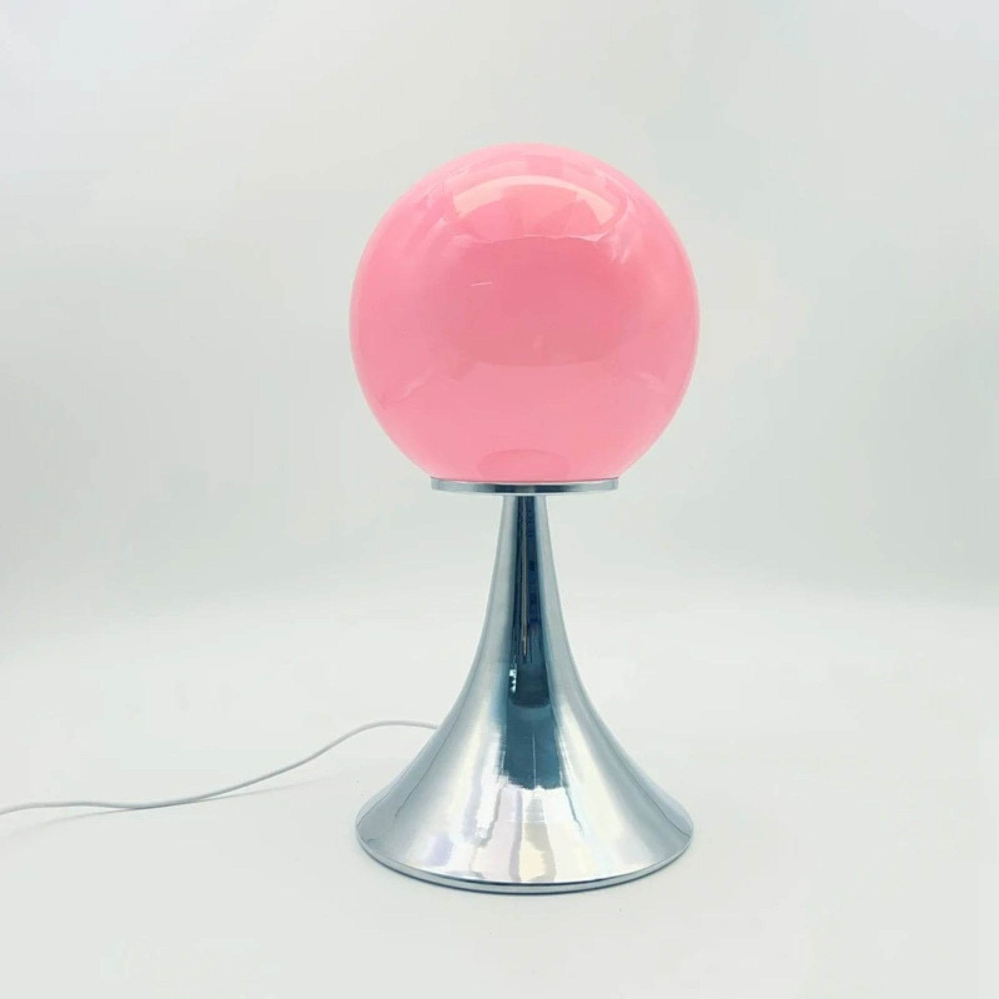 A pink glass globe table lamp on a reflective chrome base placed on a clean white tabletop.