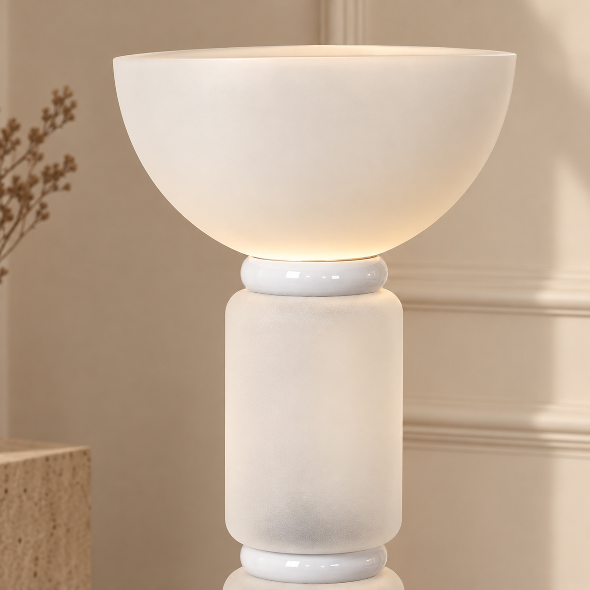 A modern table lamp with a wide, rounded bowl-shaped shade and a cylindrical base, both in a warm off-white or cream finish. The lamp is turned on, emitting a soft warm glow.
