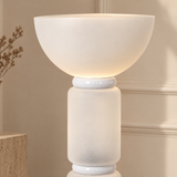 A modern table lamp with a wide, rounded bowl-shaped shade and a cylindrical base, both in a warm off-white or cream finish. The lamp is turned on, emitting a soft warm glow.