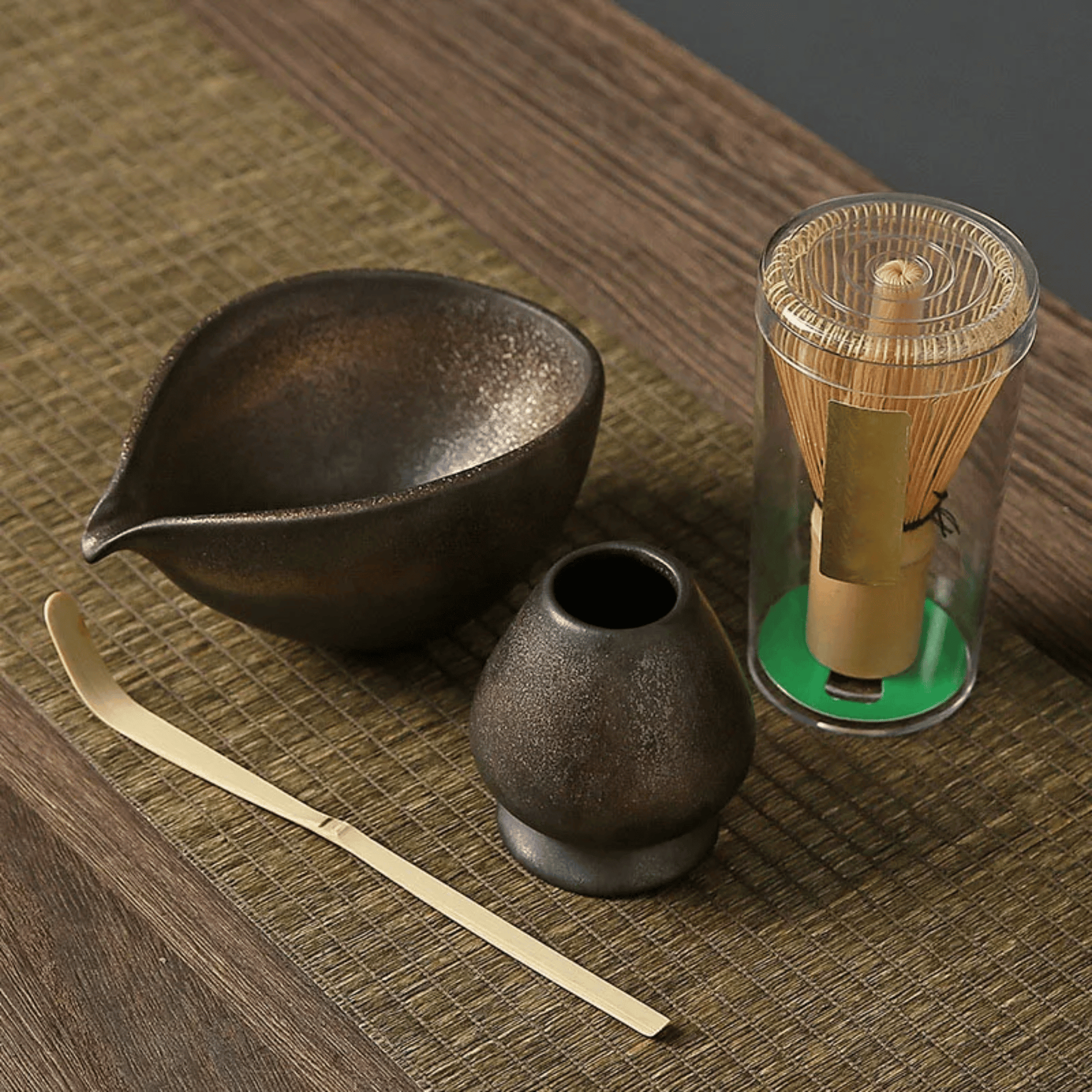 Tea-making set with black ceramic bowl, small container, gold whisk, and transparent container on a wooden surface.