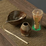 Tea-making set with ceramic bowl, whisk, scoop, and glass container on a wooden surface.