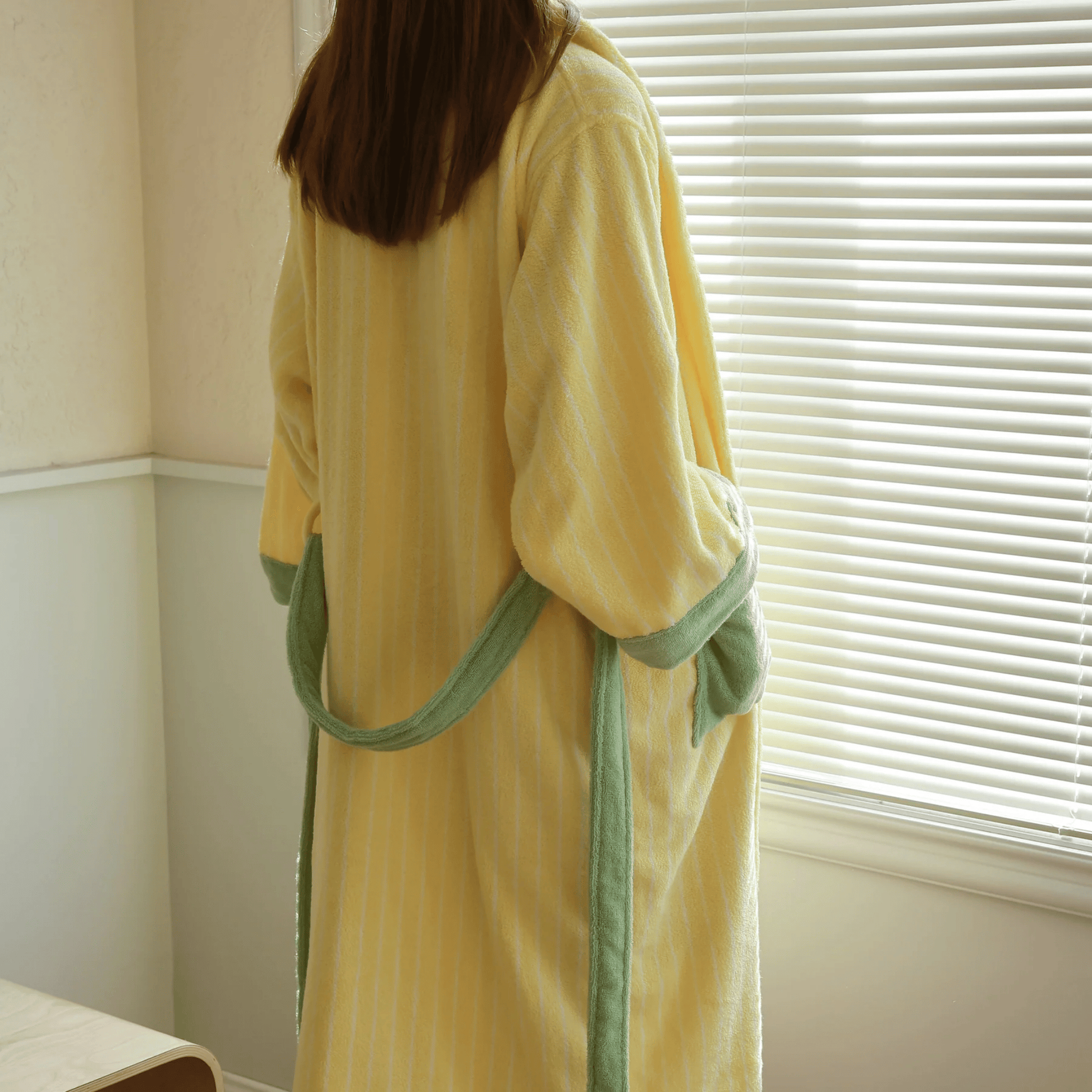 Person wearing a yellow robe with green trim standing in front of a window with blinds.