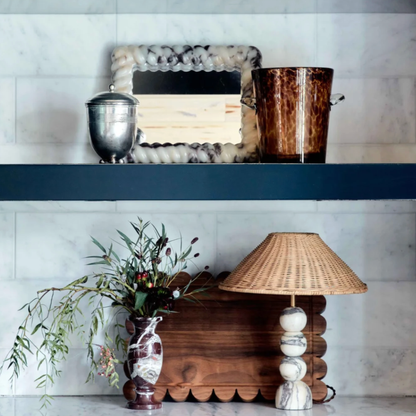 Decorative items including a mirror, urn, and vase on a shelf above a wooden table with a lamp and plant.