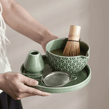 Person holding a green ceramic tea set with a bowl, whisk, and other accessories on a tray.