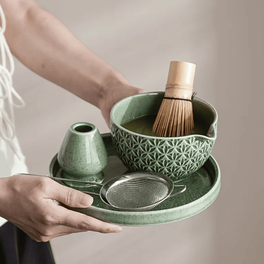 Person holding a green ceramic tea set with a bowl, whisk, and other accessories on a tray.