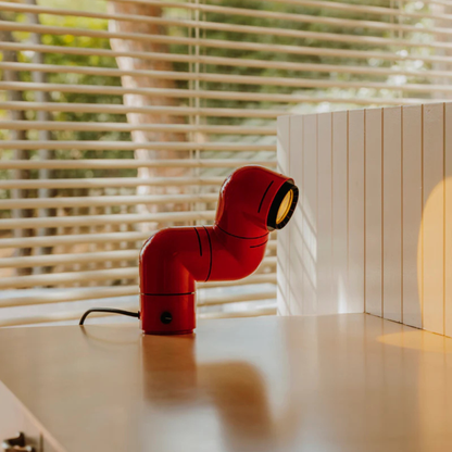 A red elbow-shaped desk lamp placed on a table with books, casting a focused beam of light in a bright setting.