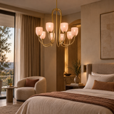 A modern multi-light chandelier with frosted glass shades hangs in a warm, elegantly styled bedroom with beige walls, a bed, and a chair.
