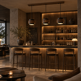 A modern bar interior with four dark wood bar stools in front of a wood counter, warm pendant lights, built-in shelving with bottles and glassware, and ambient lighting in a lounge-like setting.