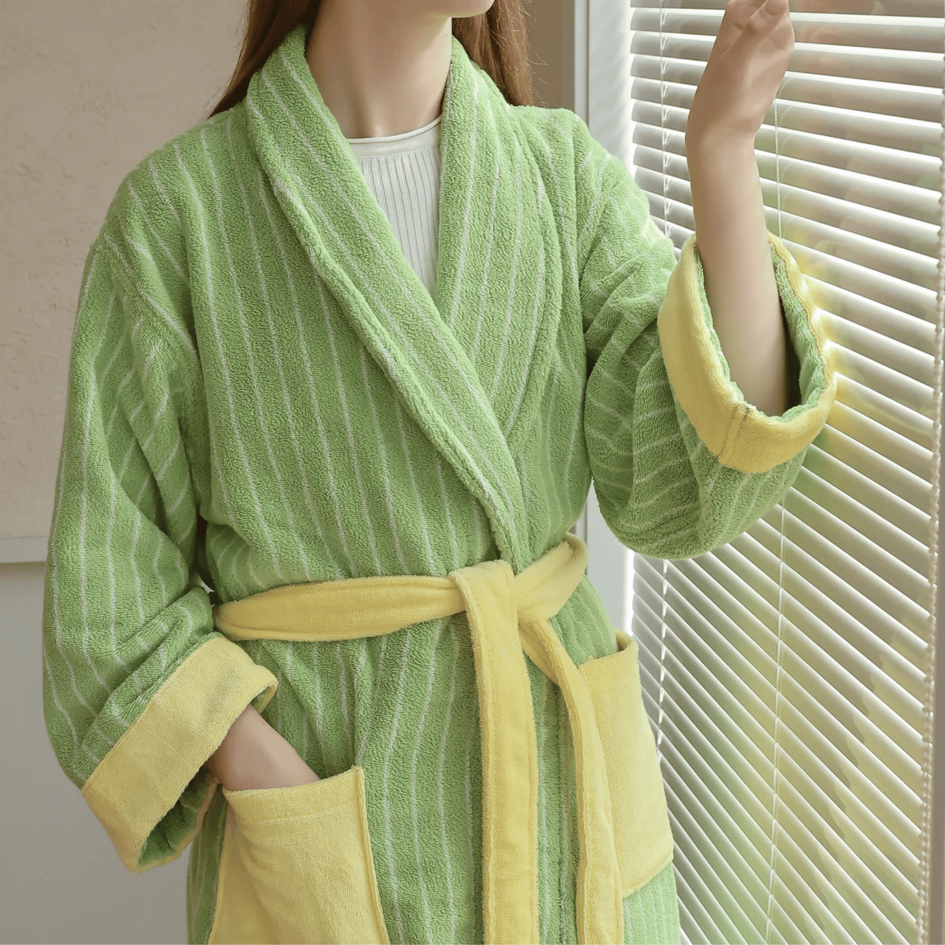 Person wearing a green and yellow robe standing by a window with blinds.