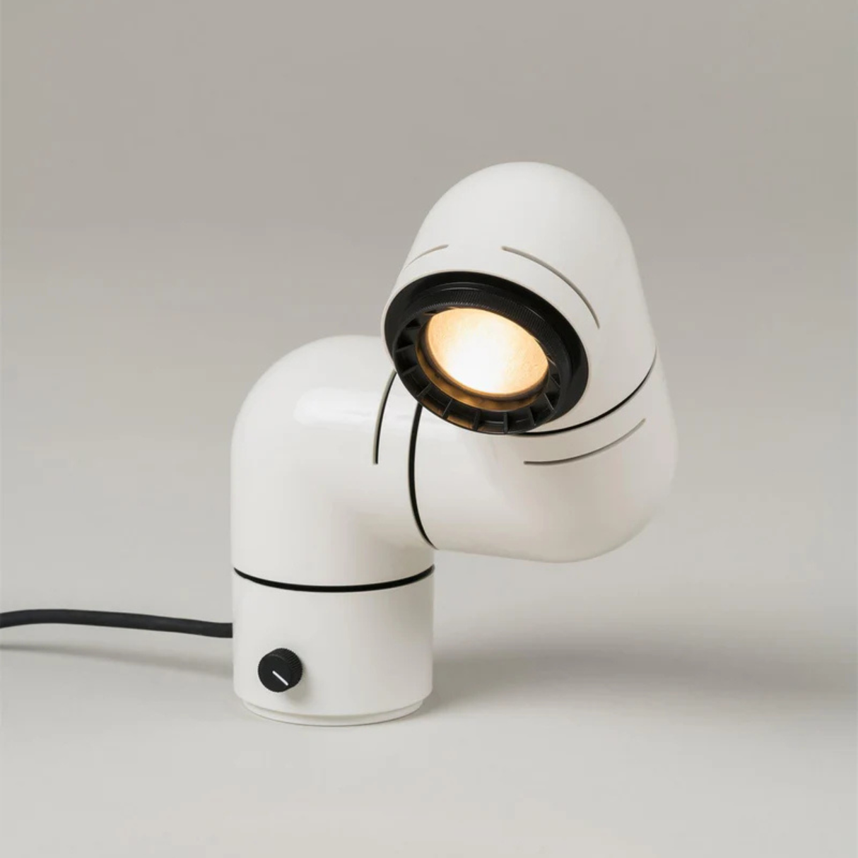 A white elbow-shaped desk lamp on a wooden table, with the light shining in a darkened room, highlighting its unique design.