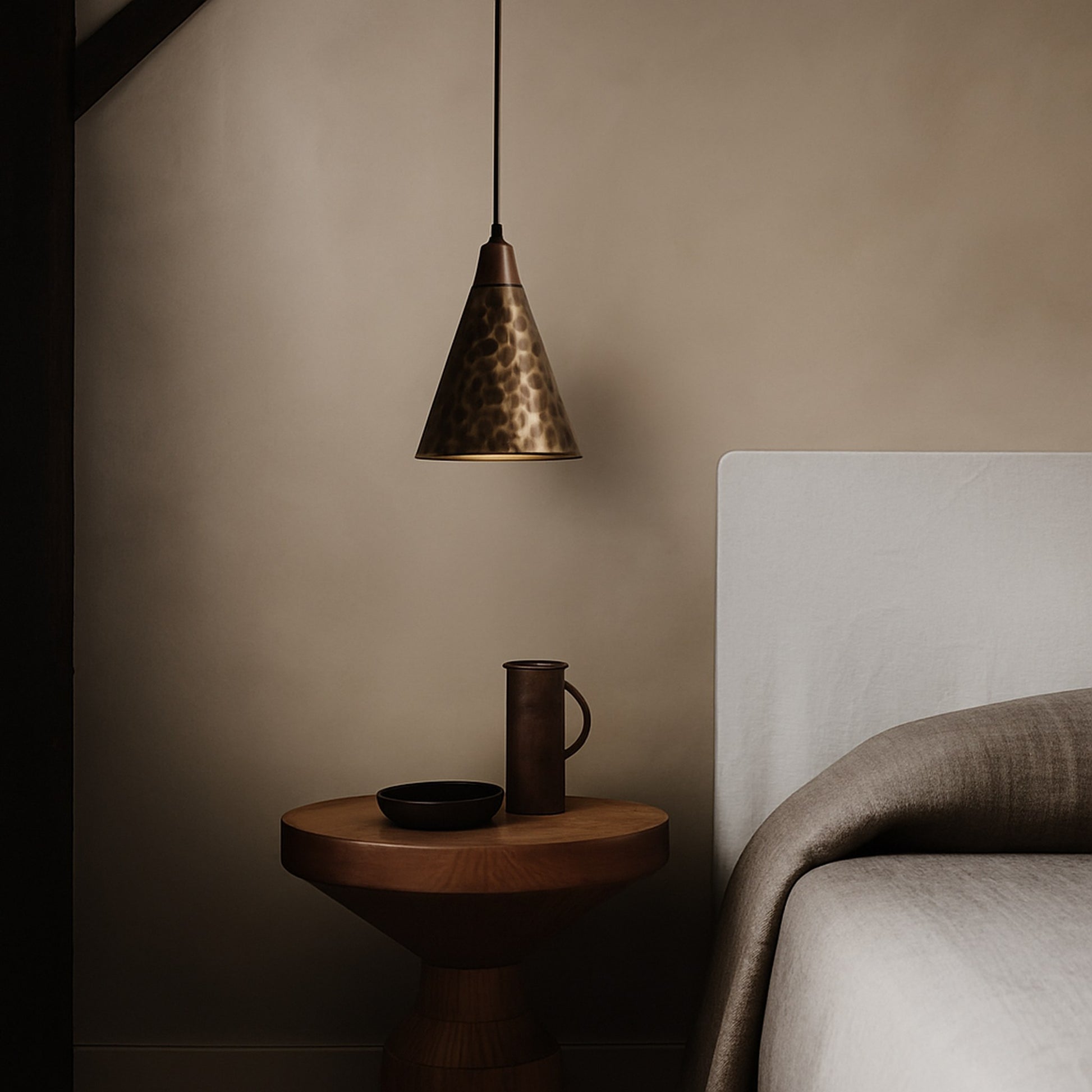 Conical pendant light above a wooden side table with a mug and bowl in a bedroom setting.