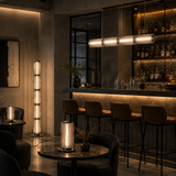 A modern bar interior with a tall cylindrical floor lamp, a matching table lamp on a round table, and a long horizontal pendant light over bar stools.