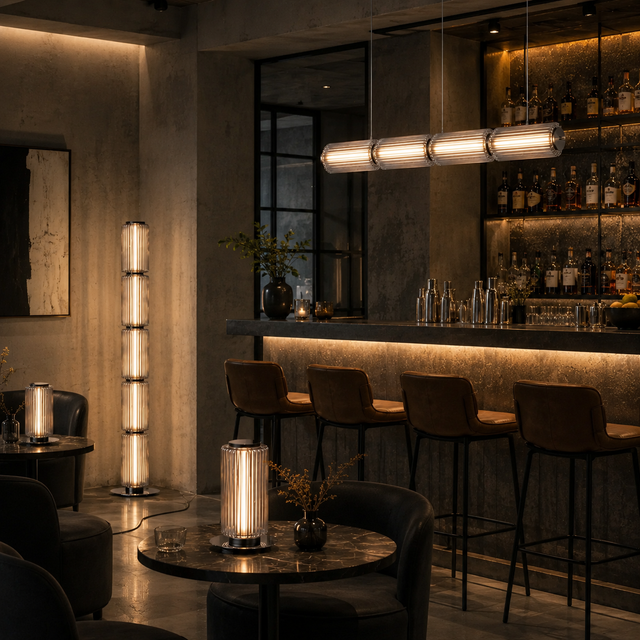 A modern bar interior with a tall cylindrical floor lamp, a matching table lamp on a round table, and a long horizontal pendant light over bar stools.