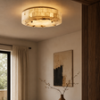 A round flush-mount ceiling light with a textured, warm-toned shell or stone-like surface, glowing in a warm beige light. It is installed on a ceiling in a modern room with beige walls, a framed abstract print, and a window with curtains.
