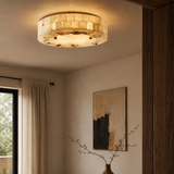 A round flush-mount ceiling light with a textured, warm-toned shell or stone-like surface, glowing in a warm beige light. It is installed on a ceiling in a modern room with beige walls, a framed abstract print, and a window with curtains.