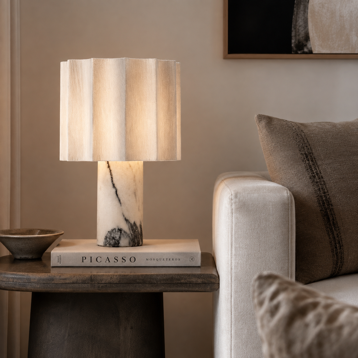 A table lamp with a pleated beige fabric shade and a white marble base with black veining, styled on a wooden side table next to a beige sofa in a warm, modern living room.